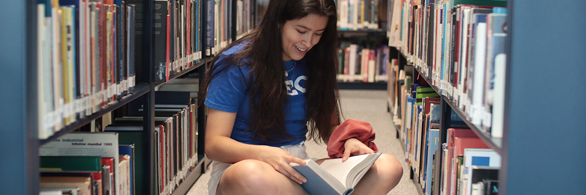 Young woman sitting in the library between bookstacks with an open book in her lap